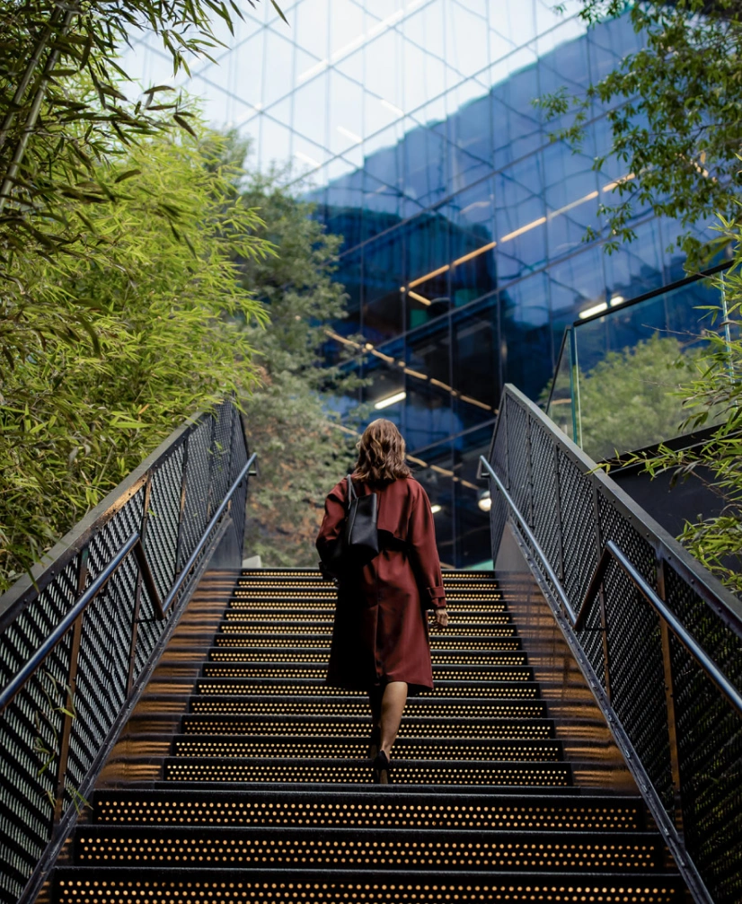 Professional woman walking up illuminated stairs toward modern glass office building surrounded by greenery