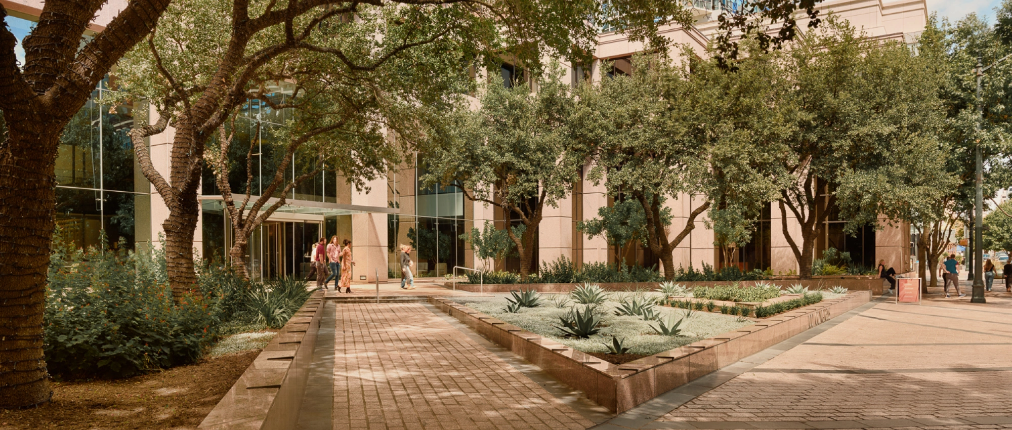 Tree shaded office plaza with landscaped walkways modern glass facade and people entering building