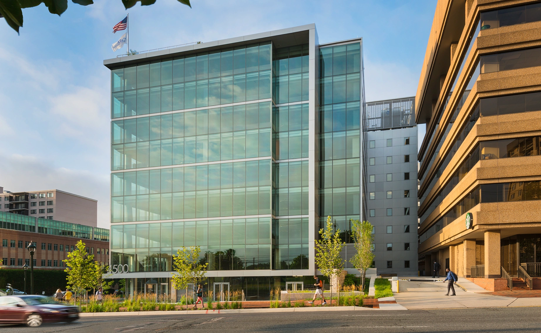 Contemporary corporate headquarters featuring glass facade, pedestrian plaza, greenery, and adjacent city architecture