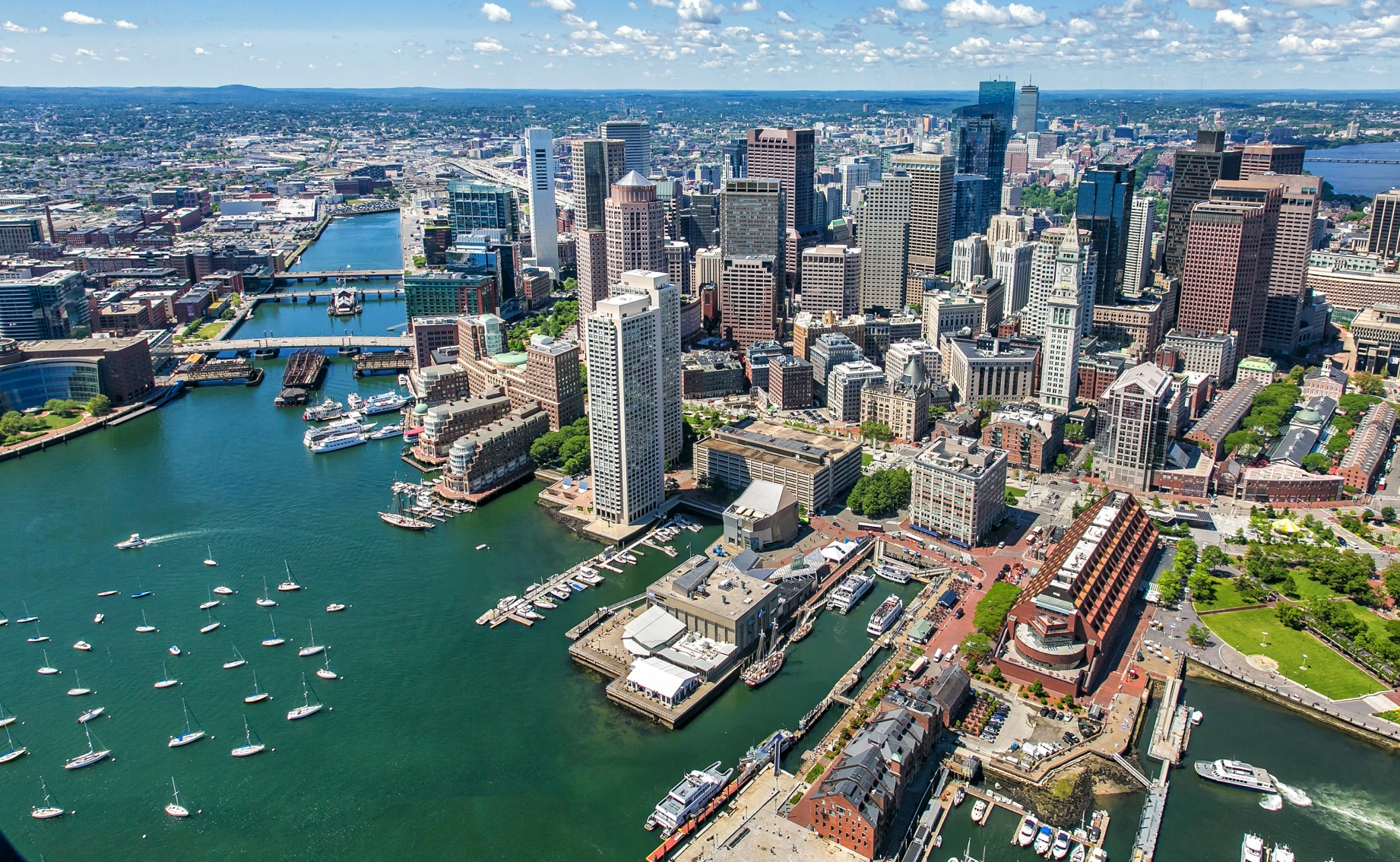 Panoramic cityscape overlooking Boston Harbor featuring high rise offices, marinas, bridges, and dense urban core