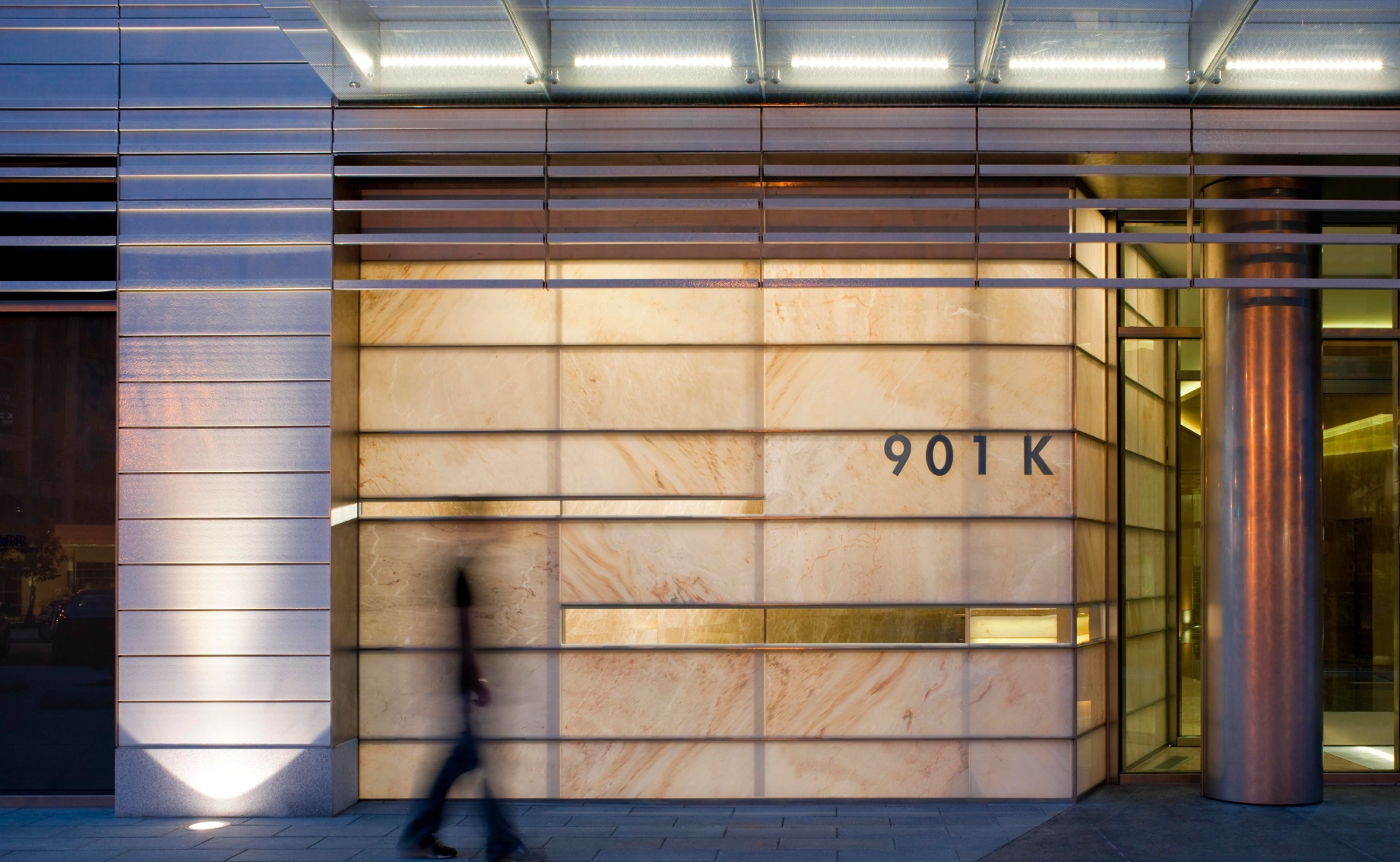 Evening streetscape highlighting a minimalist building entryway, soft architectural lighting, and a blurred pedestrian in motion