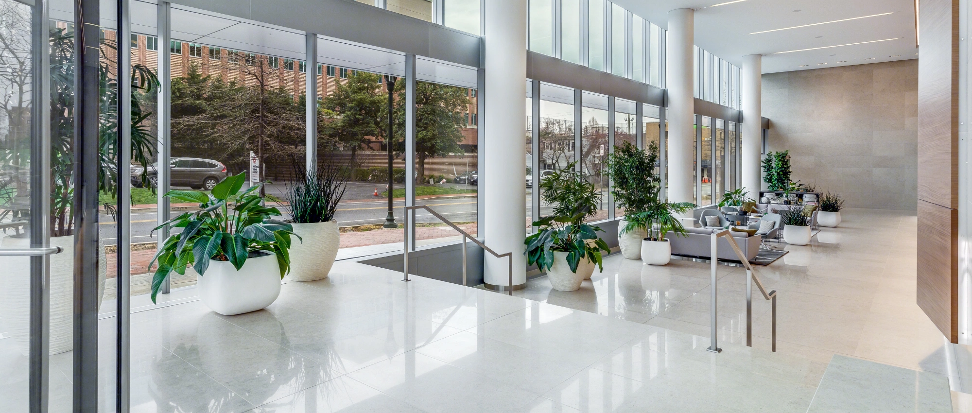Spacious office building atrium with sleek finishes, large planters, soft seating areas, and street views