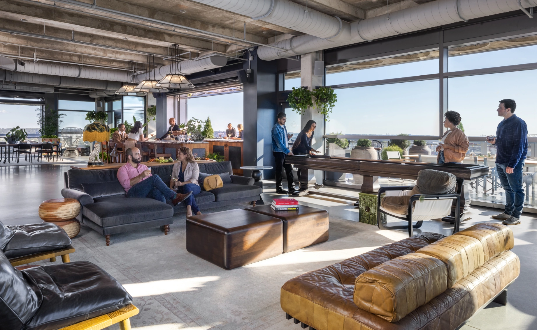 Rooftop lounge with leather seating, pool table, city views, and people socializing in sunlight