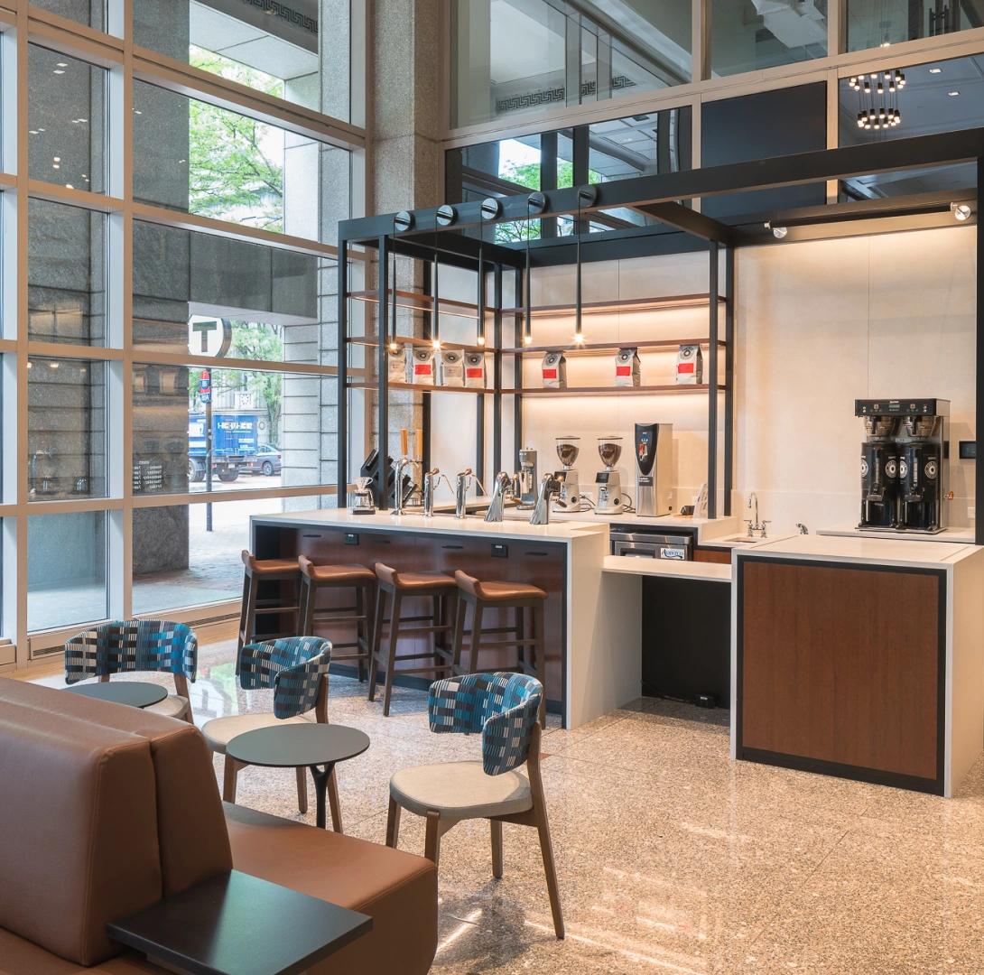 Contemporary corporate café area featuring sleek coffee counter, modern lighting, communal seating, and glass exterior walls