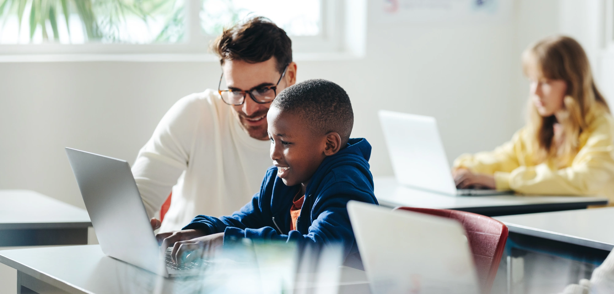 Adult educator guiding child on laptop in modern classroom with collaborative digital learning
