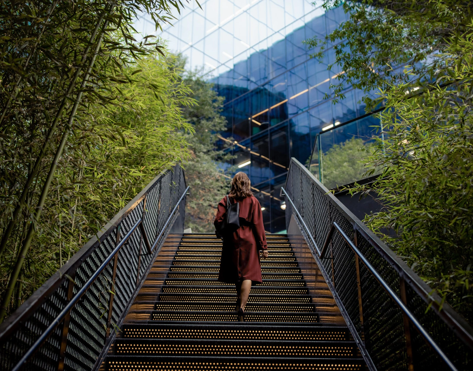 Professional woman ascending modern steps through landscaped urban space toward contemporary glass architecture