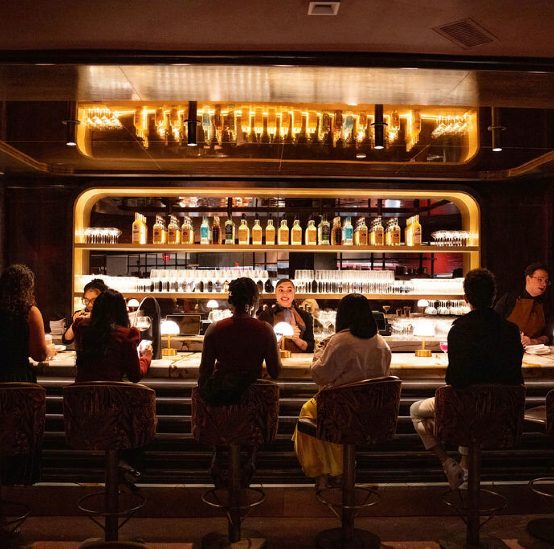 Upscale cocktail bar interior featuring illuminated shelves, bartenders serving guests, and warm ambient lighting
