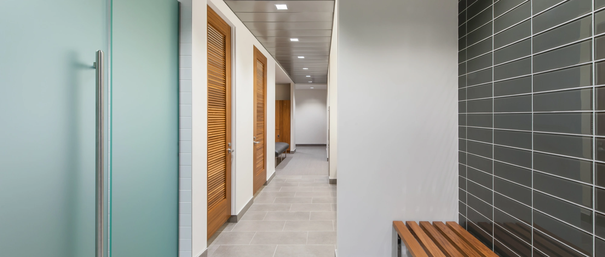 Modern locker room hallway with wood lockers, tiled walls, bench seating, and soft overhead lighting