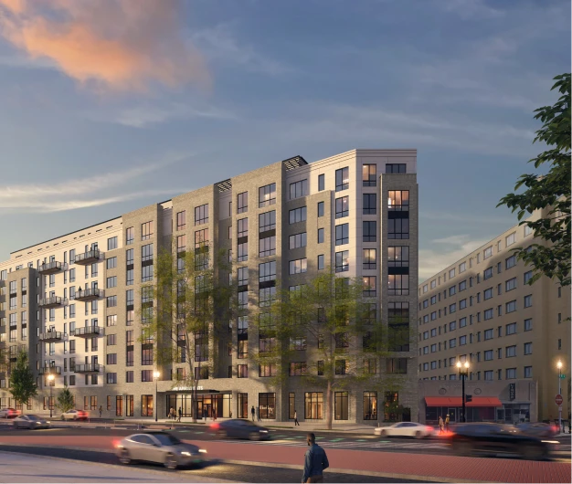 Modern mid-rise apartment building with street-level retail, trees, and city traffic at dusk
